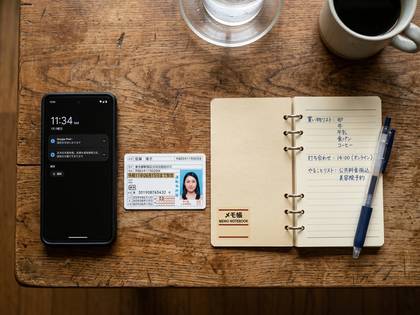 木のテーブルにスマホ、身分証、開いたメモ帳とペン。