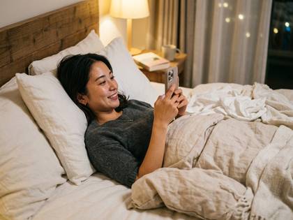 ベッドでスマホを見ながら笑顔でくつろぐアジア人女性。