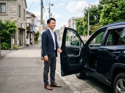 街の道路で、紺のジャケットを着た笑顔の男性が車のドアを開けている。