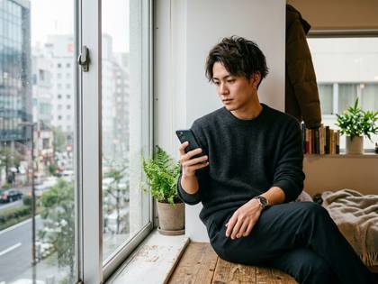 都会の窓辺に座り、スマートフォンを操作する若い男性。観葉植物も見える。