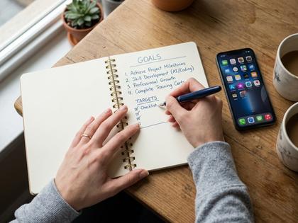 ノートに目標を書き込む女性の手元と、机に置かれたスマホの俯瞰写真