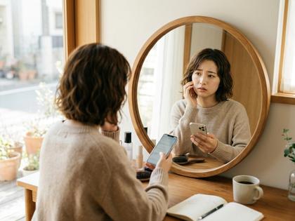 鏡を見て少し悩んだ表情でスマホを操作する女性、自然光が差し込む部屋