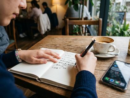 ノートに自己紹介文を下書きする女性の手元。コーヒーカップとスマホ