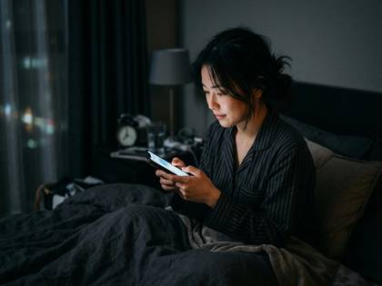 暗い寝室のベッドで、パジャマ姿の女性がスマホを操作。画面の光が顔を照らす。