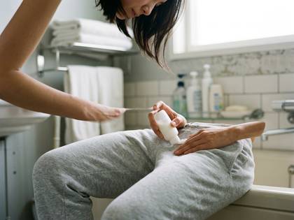 バスルームで、女性がスウェットパンツの脚にクリームを塗る。