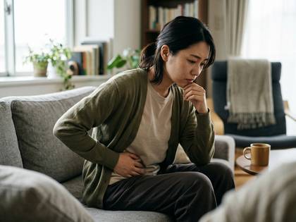 ソファに座り、腹部に手を当てて苦しむ女性。