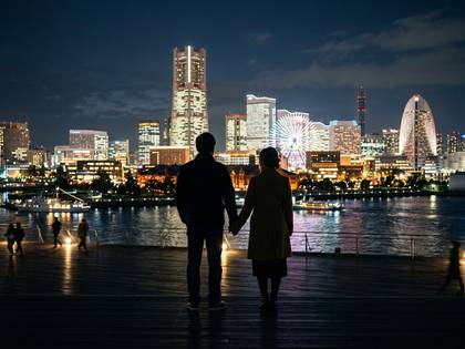 夜の水辺で手をつなぎ、きらめく街並みを見つめるカップル。