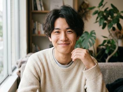 窓と本棚のある室内で笑顔を見せる若い日本人男性。