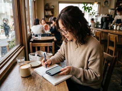 カフェで眼鏡の女性が、ノートとスマートフォンを使って作業している。