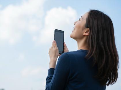 スマホを持って笑顔で空を見上げる20代女性の後ろ姿