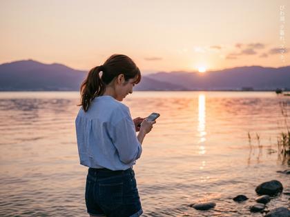 琵琶湖のほとりでスマホを見ながら微笑む20代女性の後ろ姿、夕方の柔らかな光
